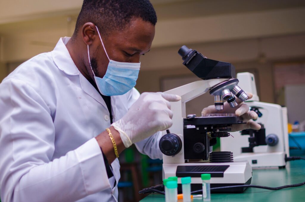 A black male scientist wearing a mask looking through a microscope.